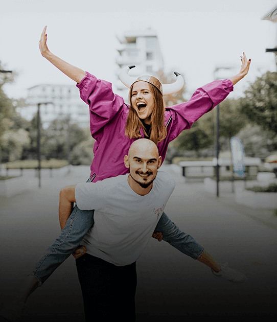 Smiling woman in a horned helmet rides on a man’s back with arms raised in joy
