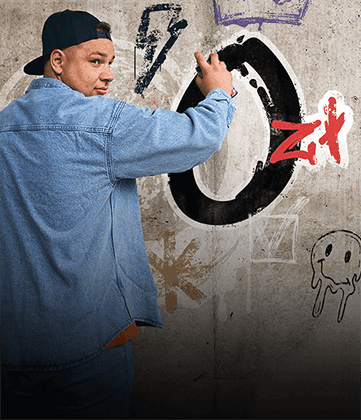 Man in a cap and denim jacket spray-paints graffiti on a concrete wall