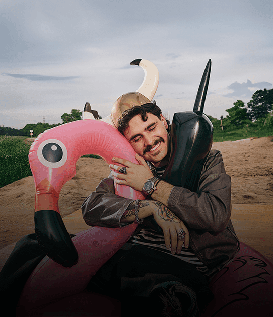 Smiling man in a horned helmet hugs inflatable flamingo and telephone against a beach backdrop