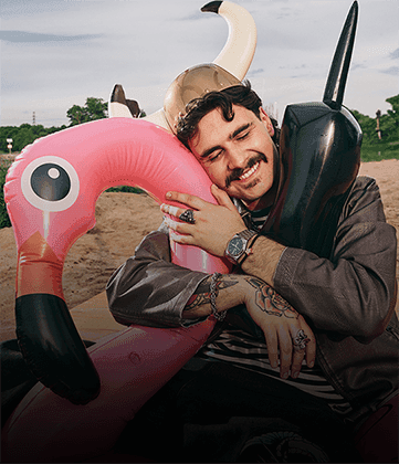 Smiling man in a horned helmet hugs inflatable flamingo and telephone against a beach backdrop
