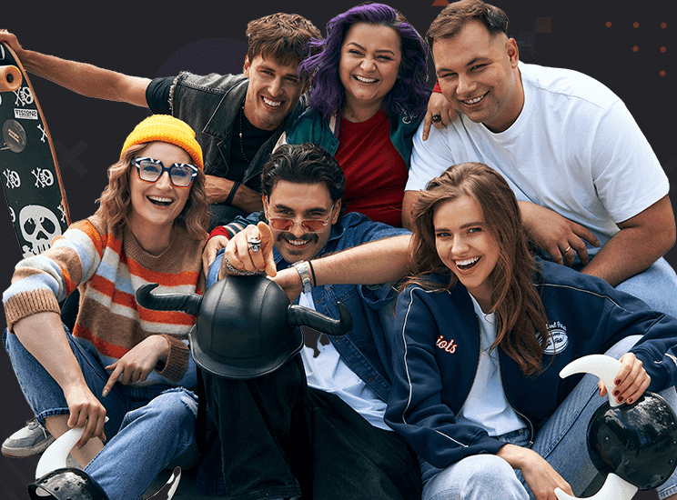 Six smiling people posing together casually; one holds a skateboard, another a horned helmet