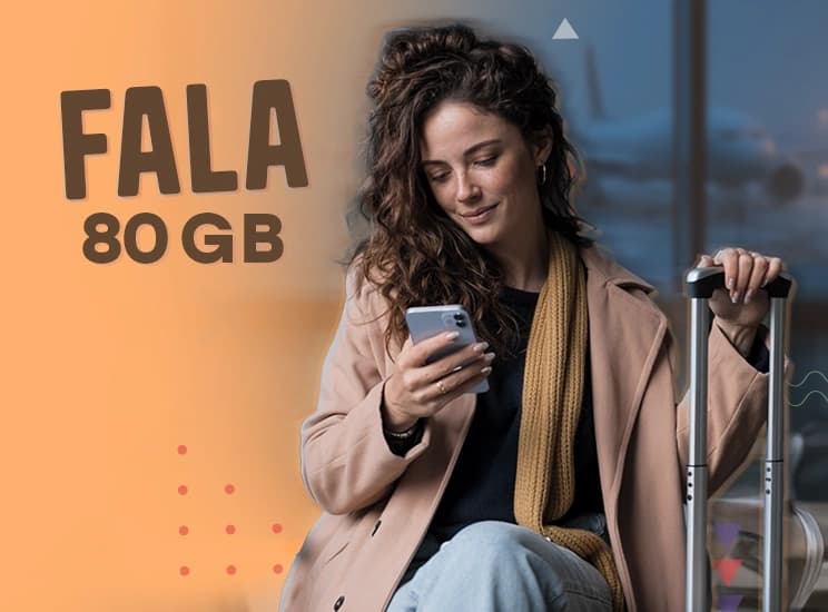 Delikatnie uśmiechnięta kobieta w płaszczu siedzi w terminalu lotniczym i spogląda na telefon trzymany jedną ręką, drugą dłoń opartą ma na wysuniętym uchwycie walizki, a w tle widać samolot stojący na pasie startowym.