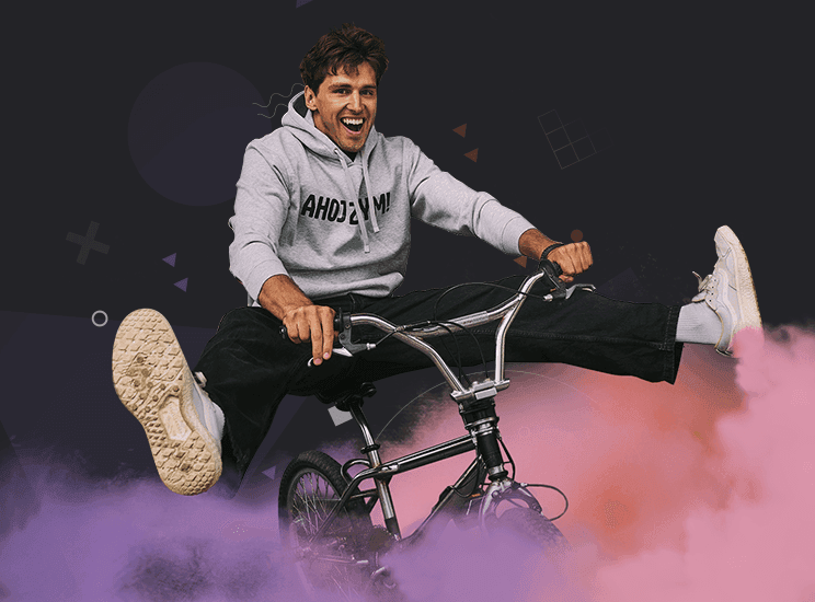 Mężczyzna w szarej bluzie jedzie na rowerze BMX z wyciągniętymi nogami, w tle kolorowy dym