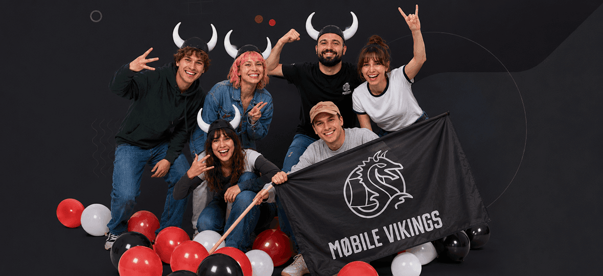 Grupa radosnych osób w hełmach z rogami pozuje wśród balonów z flagą Mobile Vikings