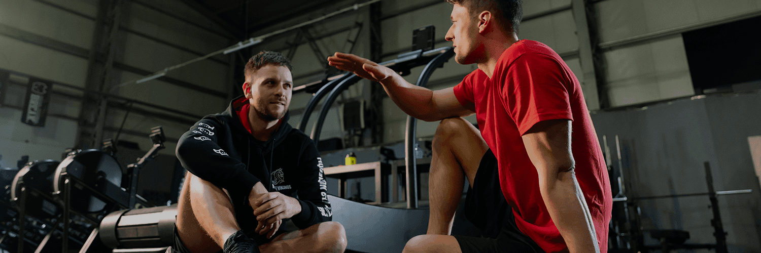 dwóch mężczyzn rozmawia w siłowni o tym, jak działa psychoedukacja. Jeden słucha, drugi żywiołowo dyskutuje