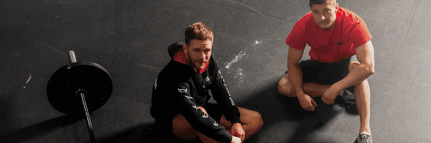 Dwóch mężczyzn na podłodze w siłowni rozmawia, widać ich z góry. Tematem rozmowy jest kryzys psychiczny.