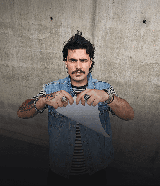 A man with a mustache and tattoos tears a sheet of paper while looking seriously at the camera